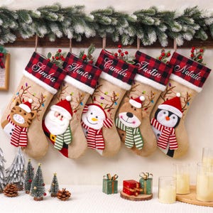 May include: Five tan burlap Christmas stockings with red and black buffalo plaid cuffs, each embroidered with a name: Simba, Hunter, Camila, Justin, and Natalie. The stockings feature plush appliques of a cat, Santa, snowman, reindeer, and penguin.