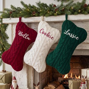 Könnte beinhalten: Drei knochenförmige Weihnachtsstrümpfe aus Zopfstrick. Der linke Strumpf ist dunkelrot mit dem in Weiß gestickten Namen Bella. Der mittlere Strumpf ist cremefarben mit dem Namen Cookie in Rot. Der rechte Strumpf ist dunkelgrün mit dem Namen Lassie in Weiß.