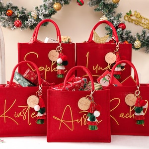 Può includere: Borse rosse con scritte dorate, ognuna personalizzata con un nome. Le borse hanno manici coordinati e portachiavi decorativi con nappe e pompon. Le borse sono piene di regali confezionati, perfette per Natale.