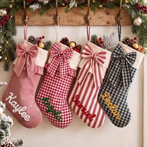 May include: Four personalised Christmas stockings hanging from a wooden rack. The first is dusty pink with the name Kaylee, the second is red and white gingham with Angela, the third is red striped with Samuel, and the fourth is navy gingham with Declan.