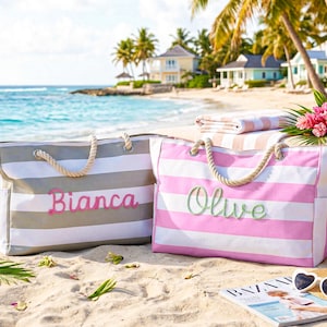 Puede incluir: Dos bolsas de playa a rayas con asas de cuerda sobre una playa arenosa. Una es gris y blanca con "Bianca" en escritura rosa. La otra es rosa y blanca con "Olive" en escritura verde. Una escena de playa con palmeras y el mar.