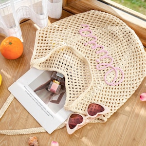 Puede incluir: Un bolso tote de ganchillo beige con un patr&oacute;n de malla abierta y la palabra "amore" bordada en letra cursiva rosa claro. El bolso contiene una tarjeta rectangular blanca con texto impreso, un frasco de perfume peque&ntilde;o y gafas de sol de coraz&oacute;n.