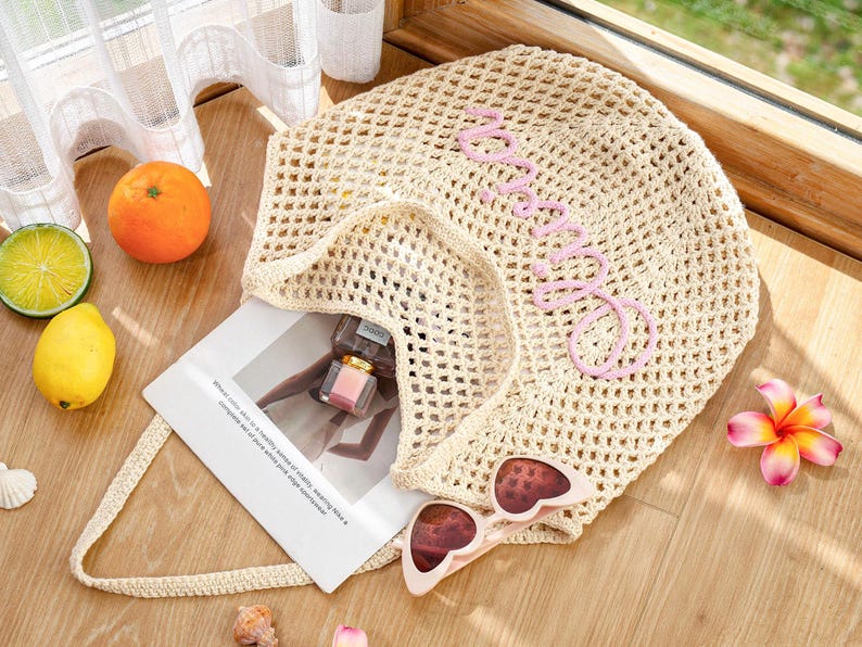 Puede incluir: Un bolso tote de ganchillo beige con un patr&oacute;n de malla abierta y la palabra "amore" bordada en letra cursiva rosa claro. El bolso contiene una tarjeta rectangular blanca con texto impreso, un frasco de perfume peque&ntilde;o y gafas de sol de coraz&oacute;n.