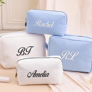 Peut inclure: Quatre trousses de maquillage personnalisées en blanc et bleu clair. Les sacs ont un motif gaufré texturé et sont brodés avec des noms ou des initiales en écriture élégante. Les sacs sont zippés et semblent être faits d'un matériau doux et durable.