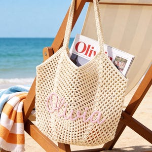 Puede incluir: Un bolso tote de ganchillo color crema con un dise&ntilde;o de malla abierta y dos correas para el hombro. La parte frontal del bolso est&aacute; personalizada con el nombre Olivia bordado en letras cursivas de color rosa claro. Una revista est&aacute; dentro.