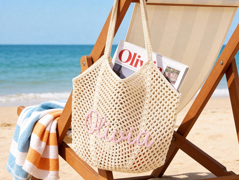 Puede incluir: Un bolso tote de ganchillo color crema con un dise&ntilde;o de malla abierta y dos correas para el hombro. La parte frontal del bolso est&aacute; personalizada con el nombre Olivia bordado en letras cursivas de color rosa claro. Una revista est&aacute; dentro.