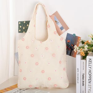 K&ouml;nnte beinhalten: Eine wei&szlig;e Einkaufstasche mit rosa Blumen Stickerei. Die Tasche hat zwei Henkel und ist aus einem weichen, strukturierten Stoff.
