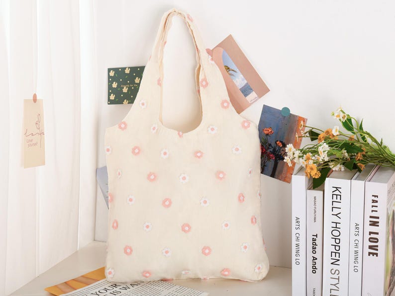 K&ouml;nnte beinhalten: Eine wei&szlig;e Einkaufstasche mit rosa Blumen Stickerei. Die Tasche hat zwei Henkel und ist aus einem weichen, strukturierten Stoff.
