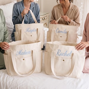 May include: Four off-white canvas tote bags with two top handles and a front pocket secured by a metal snap. Each bag features a name embroidered in light blue script: Kaylee, Camila, Madison, and Rachel. The bags have side pockets.