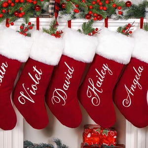 Op de afbeelding: Vijf rode kerstsokken met witte bontafwerking, elk gepersonaliseerd met een naam in elegant schrift: Justin, Violet, David, Hailey en Angela. Feestelijke vakantiedecoratie.