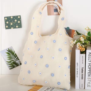 K&ouml;nnte beinhalten: Eine wei&szlig;e Einkaufstasche mit einem blauen Blumenmuster. Die Tasche hat ein schlichtes Design mit einem einzigen Henkel.