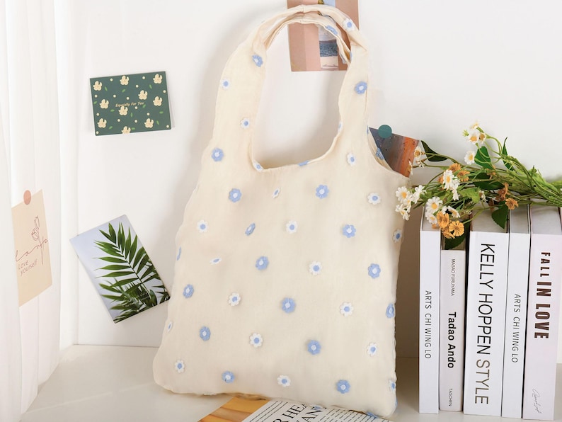 K&ouml;nnte beinhalten: Eine wei&szlig;e Einkaufstasche mit einem blauen Blumenmuster. Die Tasche hat ein schlichtes Design mit einem einzigen Henkel.