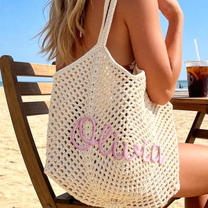 Puede incluir: Un bolso de mano de ganchillo color crema con un patr&oacute;n de malla abierta y dos correas para el hombro. La parte frontal del bolso est&aacute; decorada con el nombre Olivia escrito en letras cursivas de hilo rosa claro en relieve.