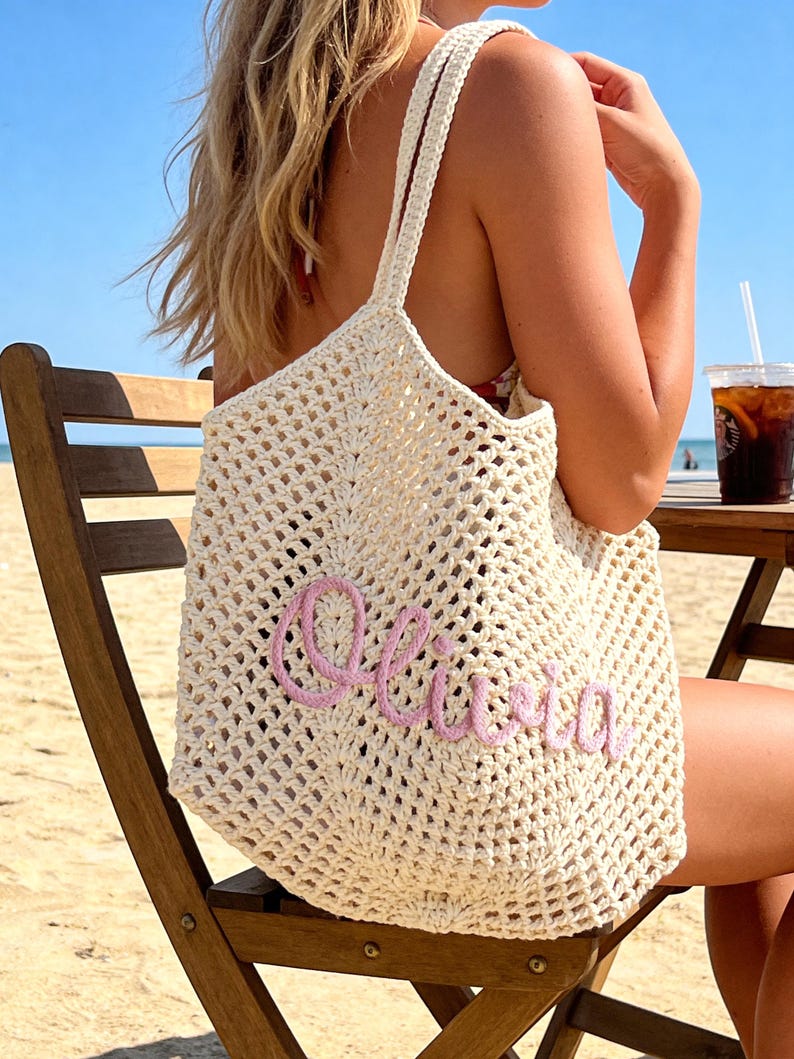 Puede incluir: Un bolso de mano de ganchillo color crema con un patr&oacute;n de malla abierta y dos correas para el hombro. La parte frontal del bolso est&aacute; decorada con el nombre Olivia escrito en letras cursivas de hilo rosa claro en relieve.