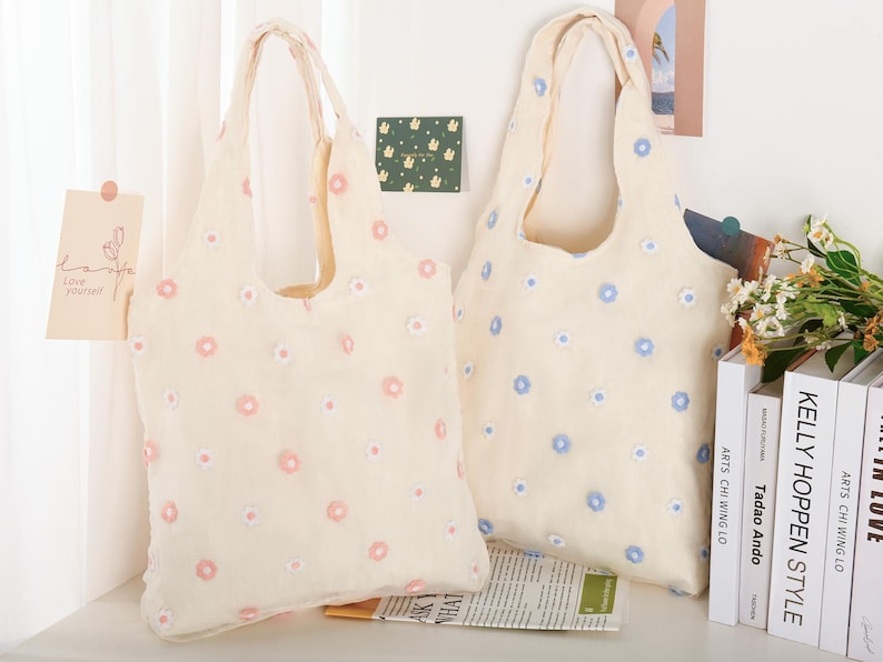 K&ouml;nnte beinhalten: Zwei beigefarbene Einkaufstaschen mit Blumenmustern. Eine Tasche hat rosa Blumen und die andere hat blaue Blumen.