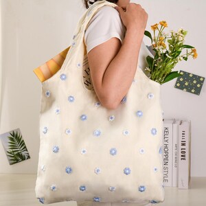 K&ouml;nnte beinhalten: Eine wei&szlig;e Tragetasche mit blauer Blumen-Stickerei. Die Tasche wird von einer Person in einem wei&szlig;en Shirt gehalten. Auf der Seite der Tasche steht "KELLY HOPPEN STYLE" gedruckt.