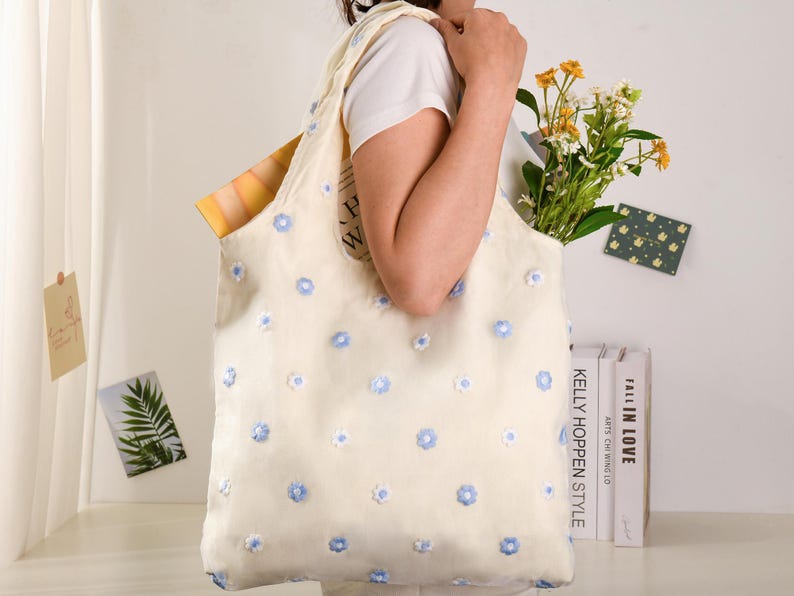 K&ouml;nnte beinhalten: Eine wei&szlig;e Tragetasche mit blauer Blumen-Stickerei. Die Tasche wird von einer Person in einem wei&szlig;en Shirt gehalten. Auf der Seite der Tasche steht "KELLY HOPPEN STYLE" gedruckt.