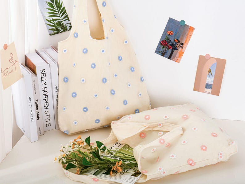 K&ouml;nnte beinhalten: Zwei beigefarbene Einkaufstaschen mit Blumenstickerei. Die gr&ouml;&szlig;ere Tasche hat blaue Blumen und die kleinere Tasche hat rosa Blumen. Beide Taschen sind aus einem leichten Stoff.