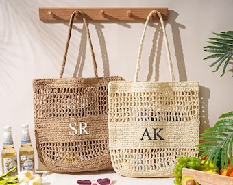 May include: Two woven tote bags with long handles hang on a wooden rack. One is light brown with the letters "SR" and the other is beige with the letters "AK". The bags have an open weave design. The scene includes drinks, fruit, and sunglasses.