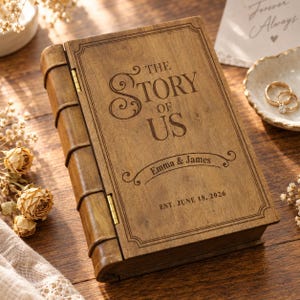 Könnte beinhalten: Eine rechteckige Holzschatulle in Buchform mit einem Klapprücken. Der Deckel ist mit dem Text THE STORY OF US, den Namen Emma & James sowie dem Datum EST. JUNE 18, 2026 graviert. Das Holz hat eine natürliche braune Maserung und Oberfläche.