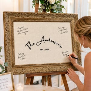 May include: A rectangular guest book sign featuring a cream-colored fabric center framed in an ornate, antique gold-colored border. The text reads The Andersons Est. 2026, surrounded by handwritten messages in black ink on the fabric surface.