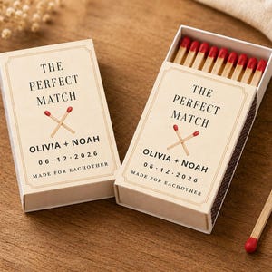 May include: Two rectangular matchboxes with cream-colored labels featuring the text THE PERFECT MATCH, OLIVIA + NOAH, 06 12 2026, and MADE FOR EACHOTHER. The labels show a graphic of two crossed matches with red tips. One box is open, revealing wooden matches.