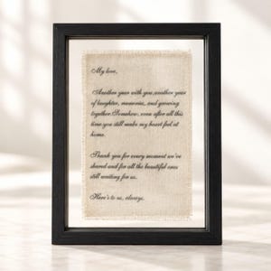 May include: A black rectangular floating frame containing a piece of cream-coloured fabric with frayed edges. The fabric features black printed text that reads: My love, Another year with you, another year of laughter, memories, and growing together.