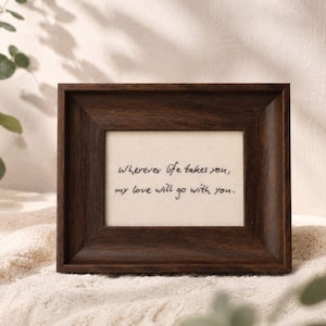 May include: A dark brown wooden picture frame containing a cream-coloured insert with the black handwritten text: Wherever life takes you, my love will go with you. The frame has a wide, beveled border and a rectangular shape, displaying a sentiment.