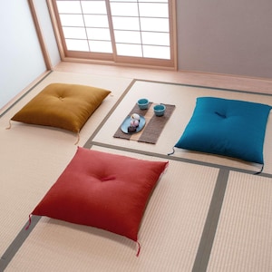 May include: Three square floor cushions in different colors: mustard yellow, teal blue, and red. The cushions are arranged on a straw mat with a small tray and two teacups in the center.