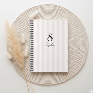 May include: A spiral-bound notebook with a striped cover featuring the letter "S" and the name "Sophia" in elegant script. The notebook rests on a beige woven placemat, with dried decorative plants to the left.