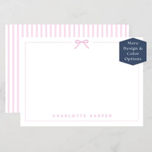 May include: A set of personalized stationery with a pink bow design. The stationery features a white background with a pink border and the name "Charlotte Harper" printed in pink. The top of the stationery has pink and white stripes. A navy blue hexagon with the text "More Design & Color Options" is on the right.