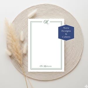 May include: White stationery with a green border and the initial "M" at the top. The text "The Madisons" is at the bottom. A navy blue hexagon with the text "More Designs & Colors" is on the right. The stationery is on a beige woven mat.