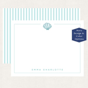 May include: A white stationery card featuring a light blue striped border, a centered light blue seashell illustration at the top, and the name EMMA CHARLOTTE printed in light blue at the bottom. A dark blue hexagon badge displays More Design & Color Options.