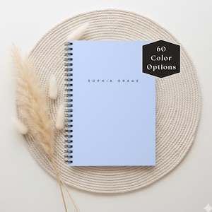 May include: A light blue spiral-bound notebook with the text "SOPHIA GRACE" in black. A black hexagon with "60 Color Options" is in the upper right corner. The notebook is on a beige woven placemat, with dried decorative plants.