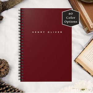 May include: A burgundy spiral-bound notebook with the text "HENRY OLIVER" in white. A black hexagon with the text "60 Color Options" is in the upper right corner. The notebook is on a white surface with other books and decorative items.