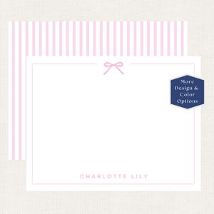 May include: A white stationery card featuring a thin pink border, a small pink bow icon at the top center, and the name CHARLOTTE LILY printed in pink at the bottom. A matching pink and white striped envelope is visible behind the card.
