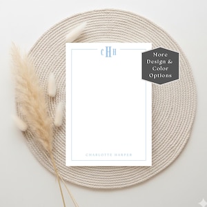 May include: A white notepad with a light blue border, monogram, and the name "Charlotte Harper" is displayed on a beige woven placemat. A sprig of dried beige pampas grass is positioned to the left of the notepad. A dark gray tag with the text "More Design & Color Options" is on the right.