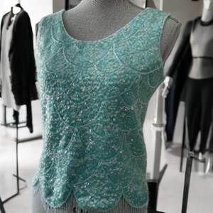 Può includere: Un top senza maniche di colore azzurro chiaro con paillettes e un orlo a balze. Le paillettes sono iridescenti e creano un effetto scintillante. Il top è su un manichino.