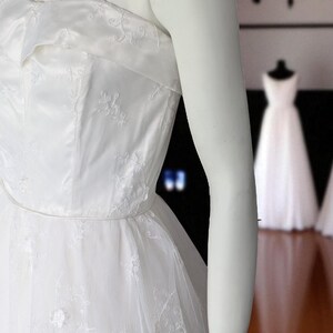 May include: A white wedding dress with a strapless bodice and delicate floral embroidery. The dress features a full, layered skirt and thin spaghetti straps. The dress is displayed on a mannequin.