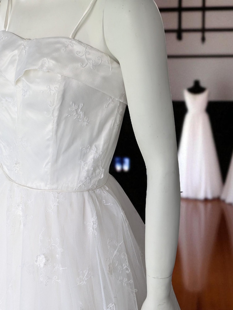 May include: A white wedding dress with a strapless bodice and delicate floral embroidery. The dress features a full, layered skirt and thin spaghetti straps. The dress is displayed on a mannequin.