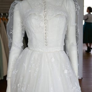 May include: A white bridal gown with a high, buttoned collar and long sleeves. The dress is embellished with floral embroidery and has a full, layered skirt. The mannequin wears a pearl choker. The dress is a classic design, suitable for a wedding.