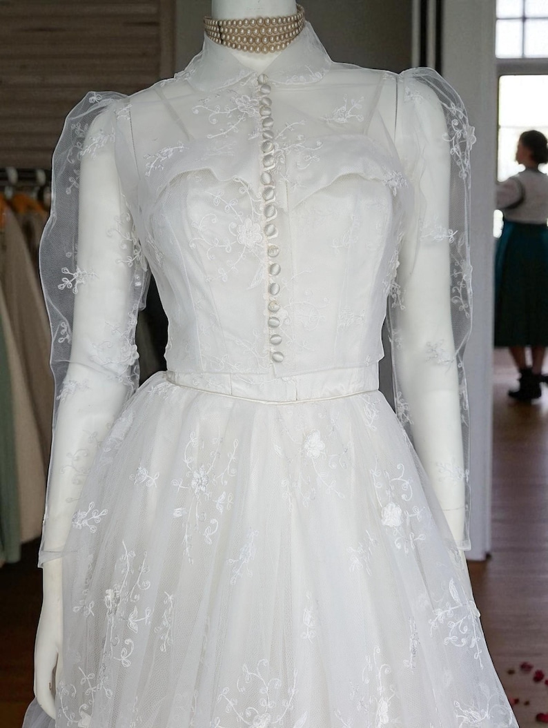 May include: A white bridal gown with a high, buttoned collar and long sleeves. The dress is embellished with floral embroidery and has a full, layered skirt. The mannequin wears a pearl choker. The dress is a classic design, suitable for a wedding.