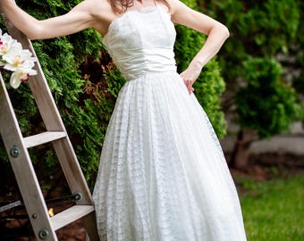 May include: A white lace dress with a strapless neckline and a tea-length skirt. The dress features a fitted bodice and a full, flowing skirt. The woman is standing next to a wooden ladder with a bouquet of white flowers.