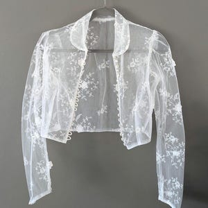 May include: A sheer white bolero jacket with long sleeves and a collar, hanging on a silver hanger. The jacket is decorated with delicate floral embroidery. The fabric appears lightweight and airy, ideal for layering.