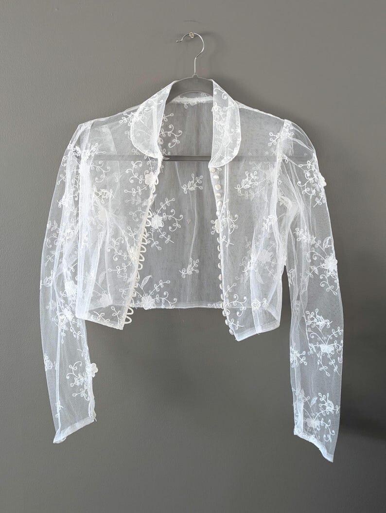 May include: A sheer white bolero jacket with long sleeves and a collar, hanging on a silver hanger. The jacket is decorated with delicate floral embroidery. The fabric appears lightweight and airy, ideal for layering.