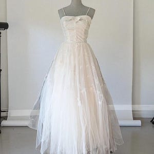 May include: An ivory wedding dress displayed on a mannequin. The dress has a sweetheart neckline, thin spaghetti straps, and a bow detail. The fitted bodice leads to a full skirt made of layered tulle. The dress is a classic, elegant style, perfect for a wedding.