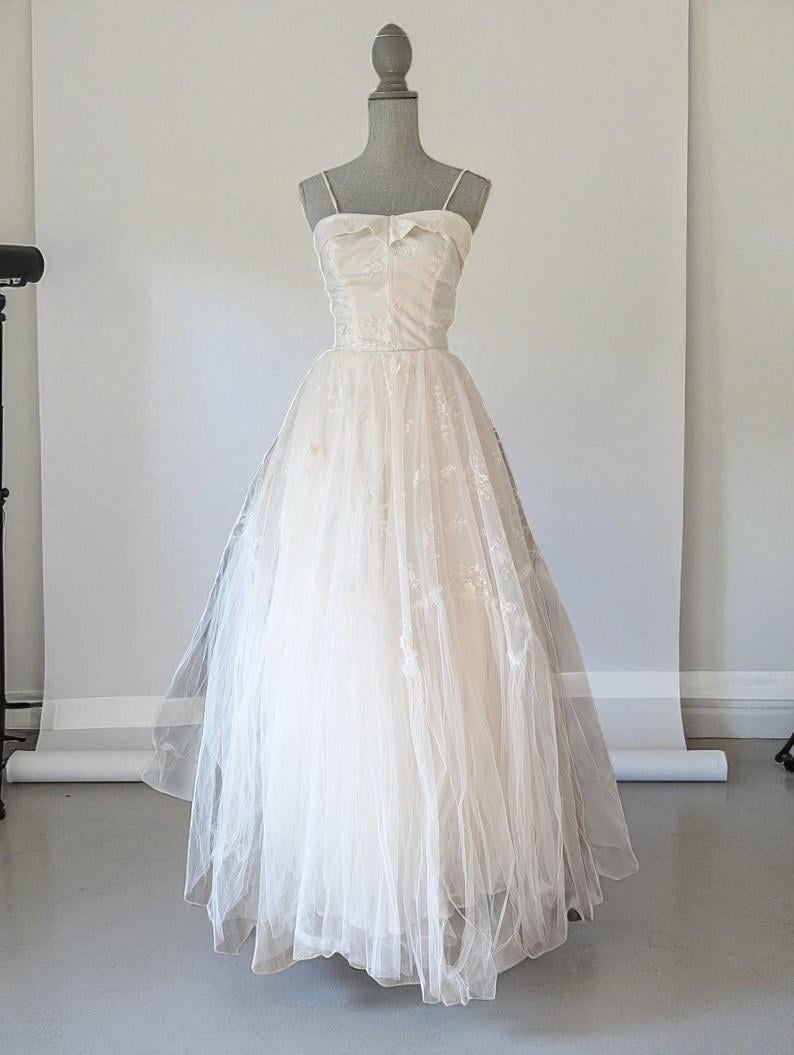 May include: An ivory wedding dress displayed on a mannequin. The dress has a sweetheart neckline, thin spaghetti straps, and a bow detail. The fitted bodice leads to a full skirt made of layered tulle. The dress is a classic, elegant style, perfect for a wedding.