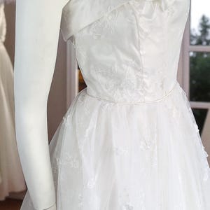 May include: A white wedding dress with a strapless neckline and a layered tulle skirt. The bodice features delicate floral embroidery. The dress is displayed on a mannequin, showcasing its elegant design and details.