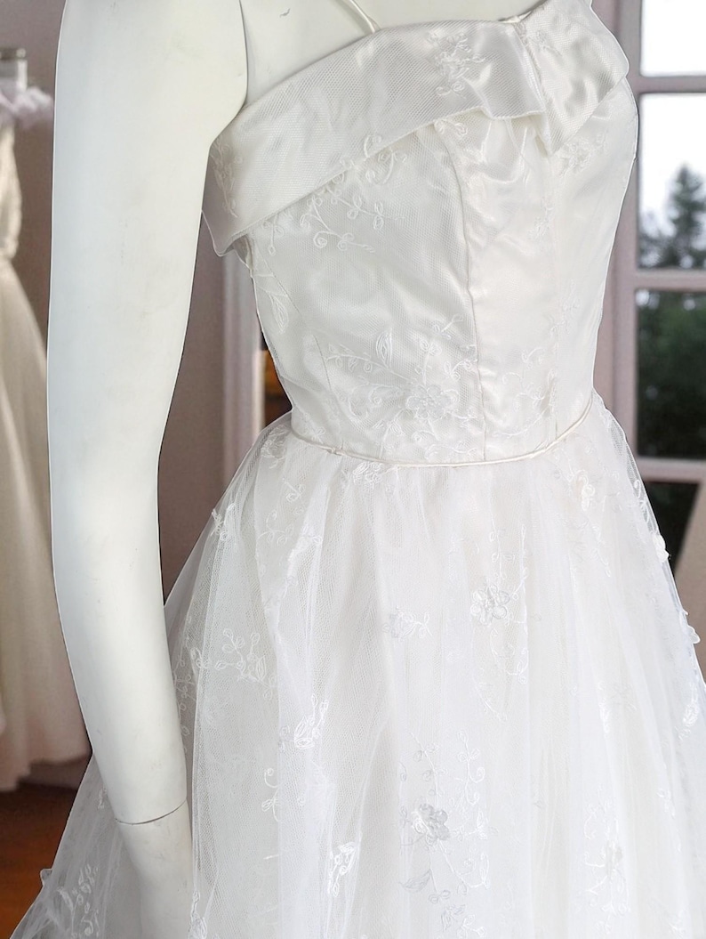 May include: A white wedding dress with a strapless neckline and a layered tulle skirt. The bodice features delicate floral embroidery. The dress is displayed on a mannequin, showcasing its elegant design and details.