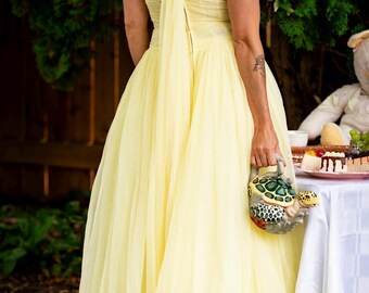 May include: A pale yellow, vintage-style dress with a flowing skirt and off-the-shoulder neckline. The dress is paired with a turtle-shaped handbag. The scene is set outdoors, with a table in the background laden with desserts.